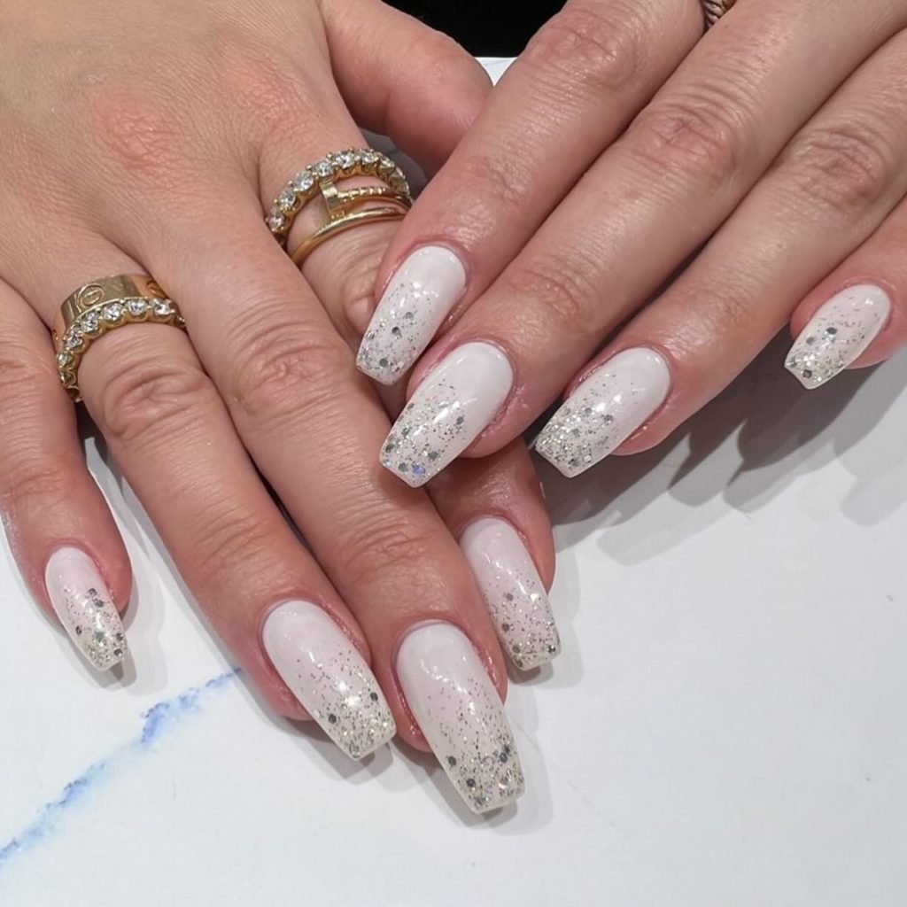 Elegant white and glitter nails with gold rings on hands, showcasing chic manicure and jewelry style.