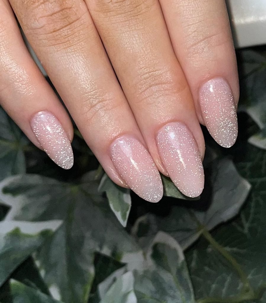Glittery nude almond-shaped nails on a leafy background, showcasing elegant and stylish nail art design.