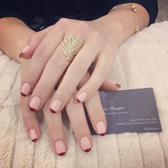 Elegant nude nails with red tips and a statement ring showcase chic nail art, resting on a luxurious fabric.