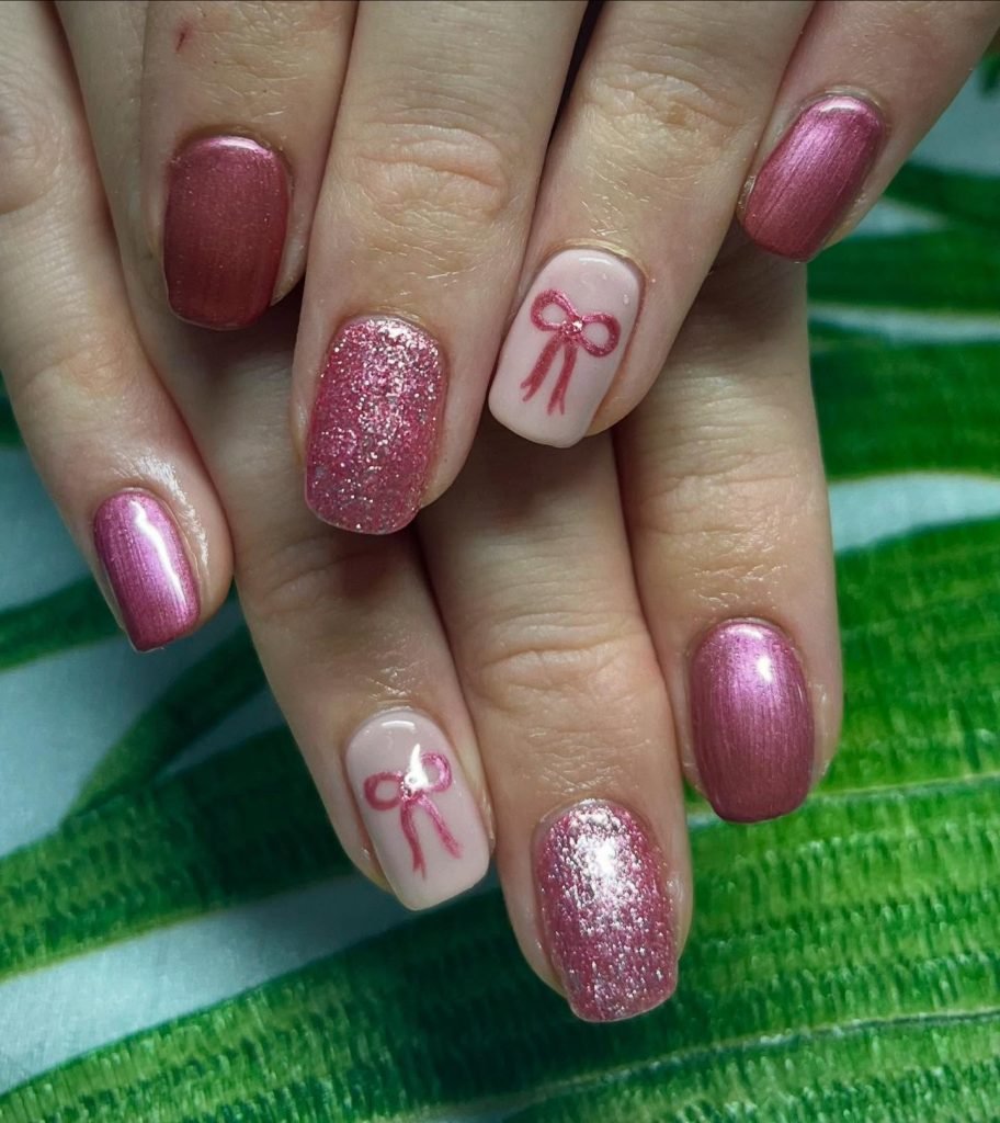 Pink and glitter manicure with bow accents on nails, displayed on green textile background. Fashionable nail art design.