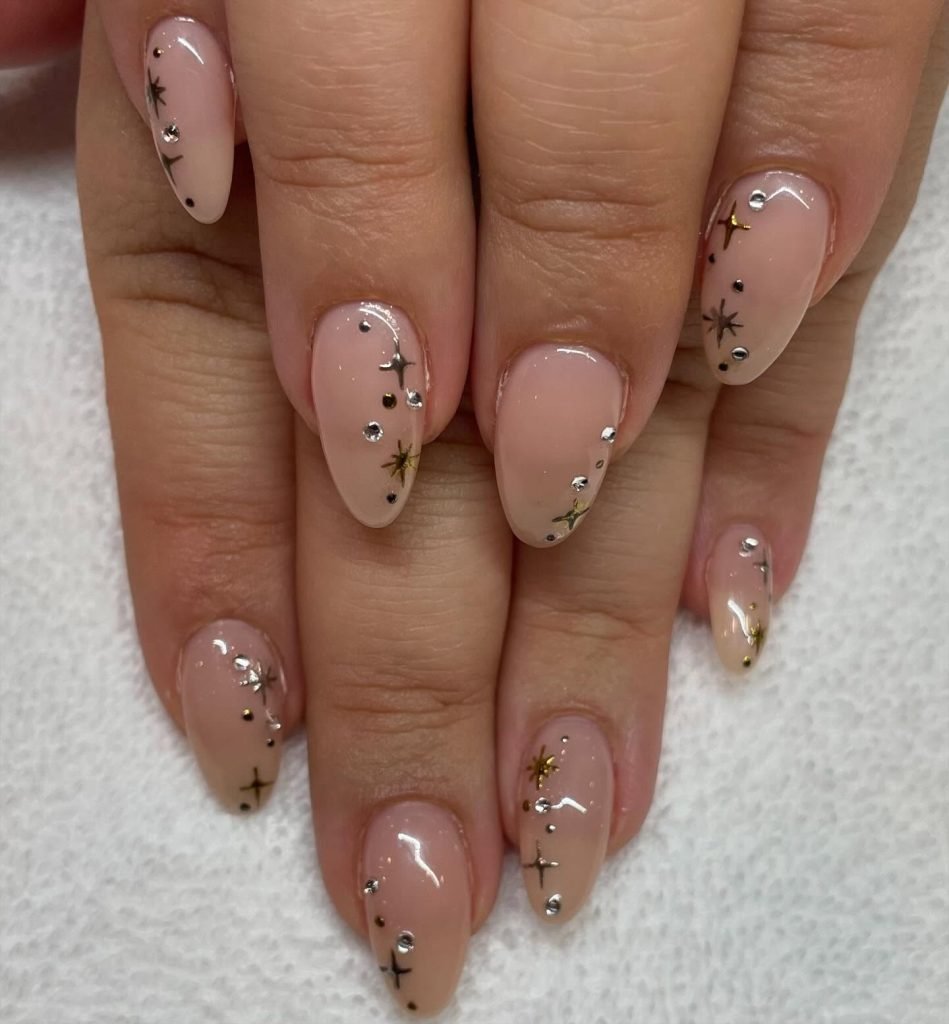 Almond-shaped nails with nude polish, adorned with silver rhinestones and gold star accents, on a white background.