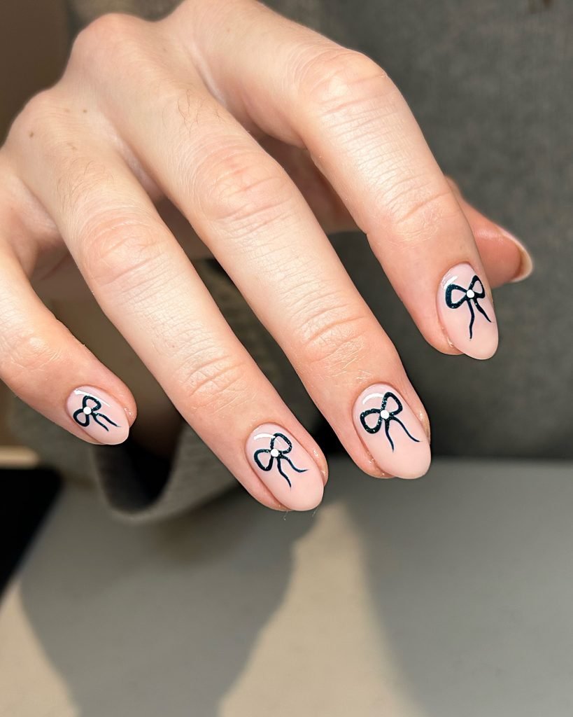 Nails painted with a nude polish featuring delicate black bow designs. Stylish and elegant nail art close-up.