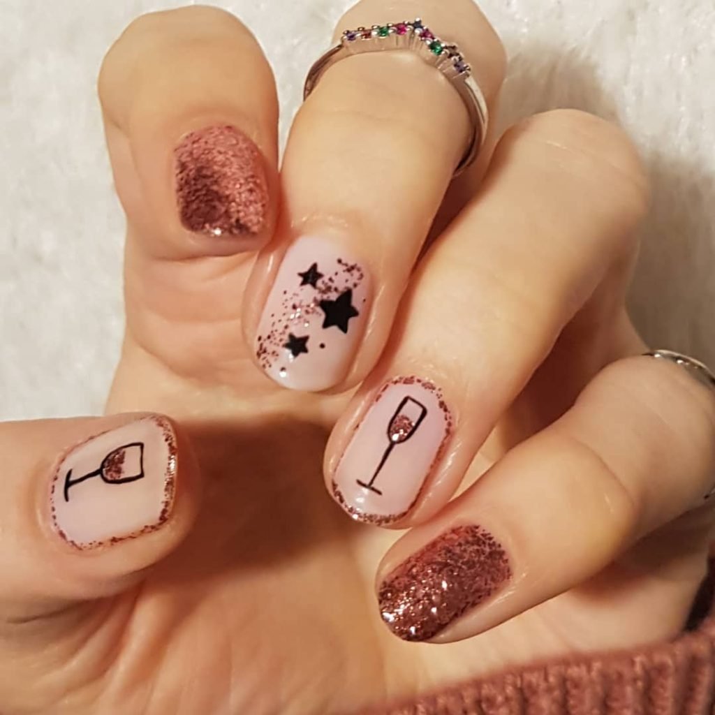 Chic nail art with rose gold glitter, star designs, and wine glass motifs on a hand wearing a colorful gemstone ring.