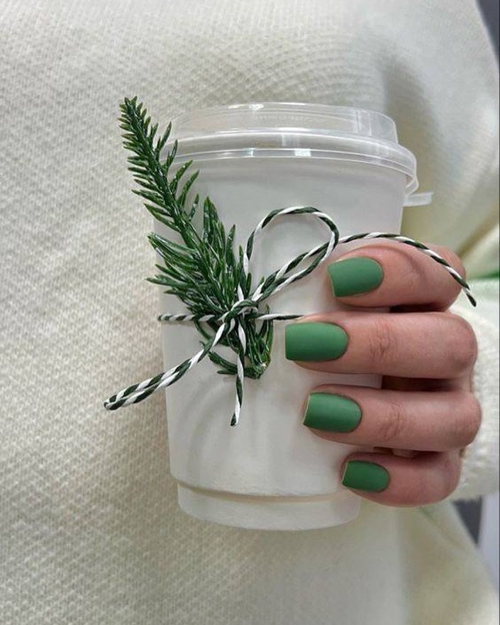 Hand with green nails holding a festive cup adorned with a pine sprig and twine, against a cozy background.