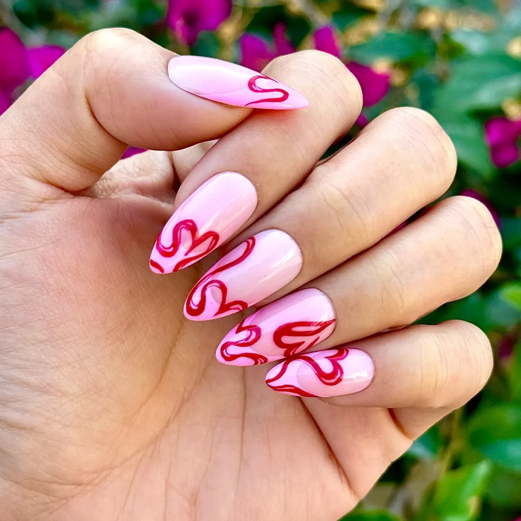 Pink nails with a glossy finish featuring red abstract swirl designs, held against a green and purple floral background.