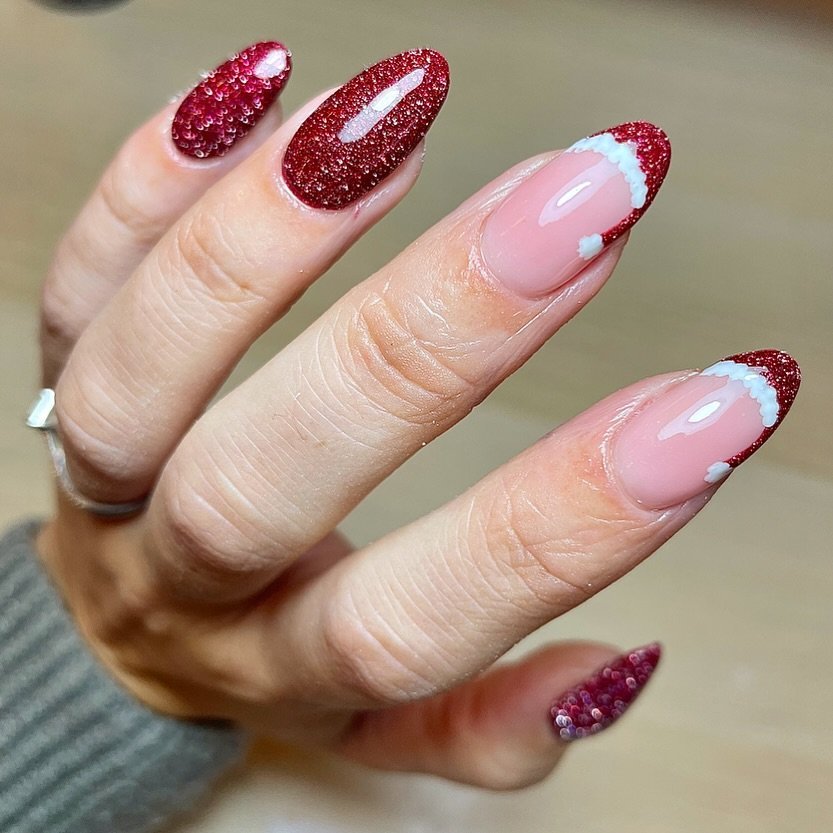 Festive red glitter nails with Santa hat tips on a manicured hand, showcasing holiday nail art design.