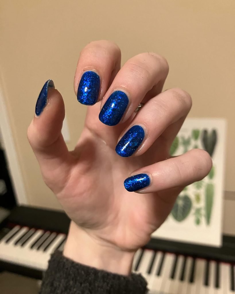 Blue glittery nail polish on hand with keyboard and plant artwork in background.
