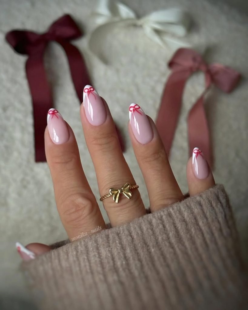 Elegant nude nails with red bow design, hand wearing a gold bow ring, set against a soft backdrop with ribbons.