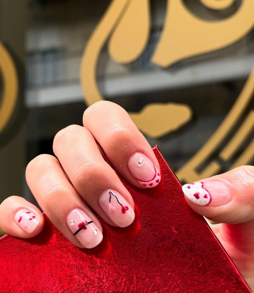 Chic Valentine's Day nail art featuring red heart designs and crystals on a hand holding a red textured surface.