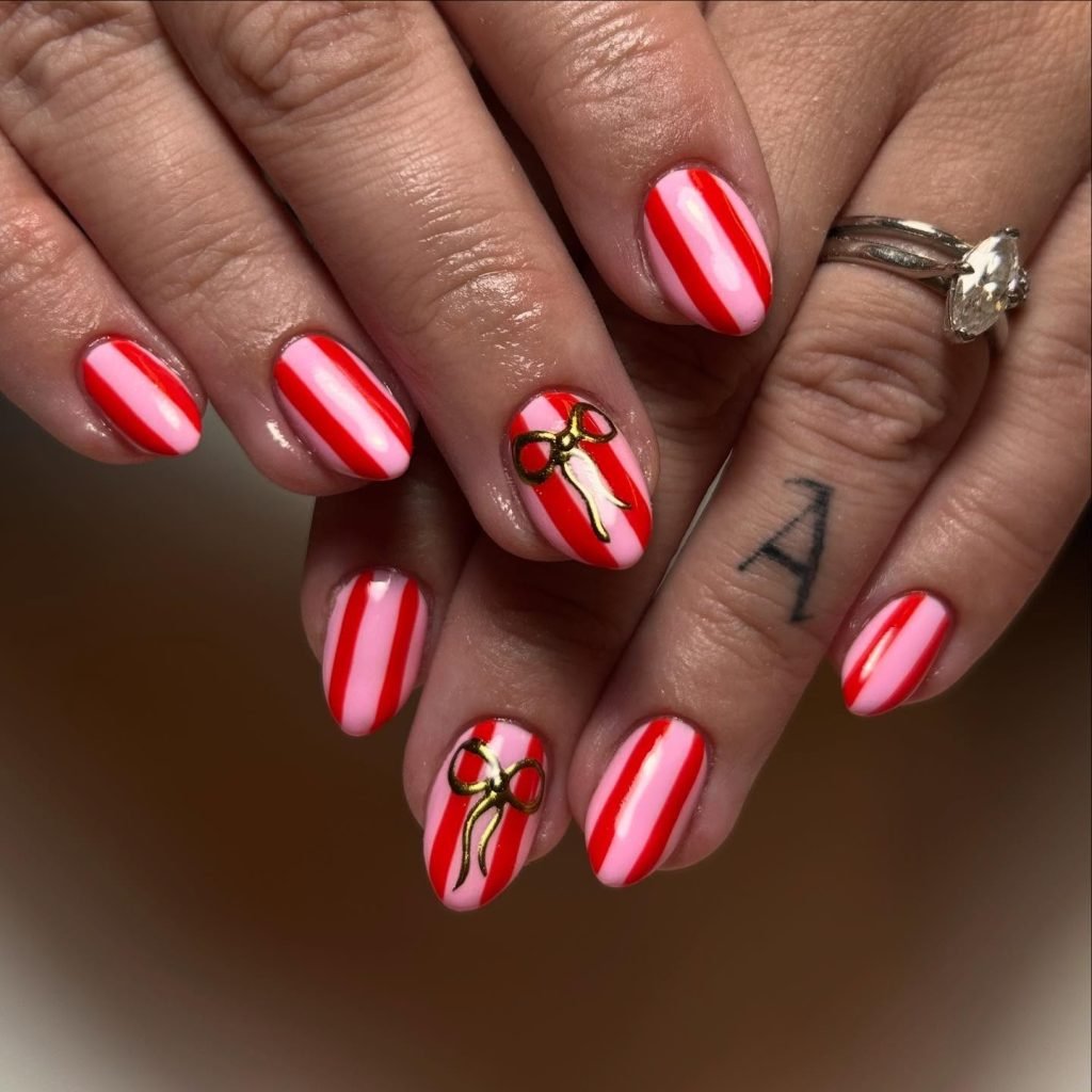 Red and pink striped nails with gold bow accents, close-up on hands with rings and a letter tattoo on finger.