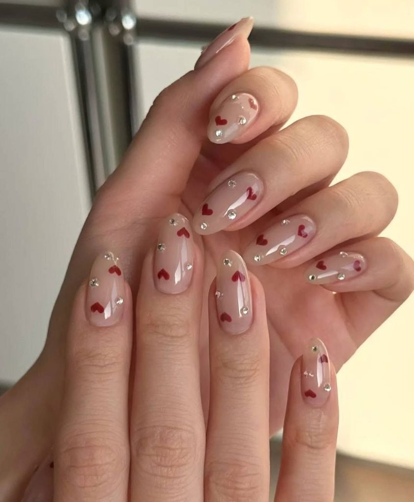 Nail art with red heart design and rhinestones on natural nails, perfect for a romantic and elegant look.