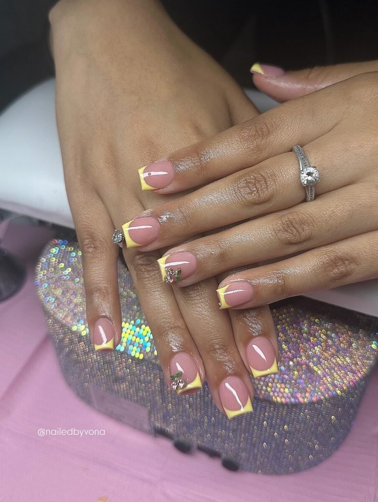Nail art featuring yellow French tips with rhinestones and floral accents on manicured hands with rings and a sparkly clutch.