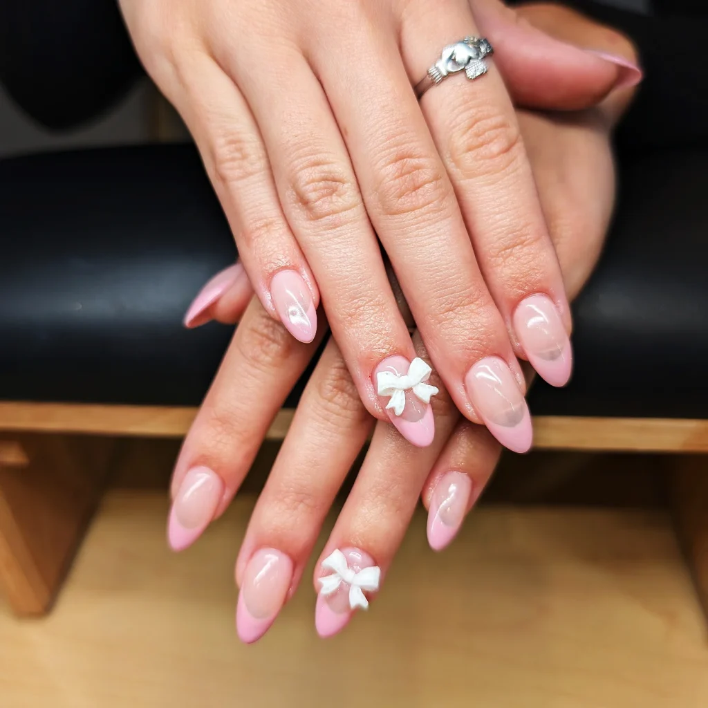 Close-up of elegant pink almond nails with white 3D bows and a silver ring on fingers.