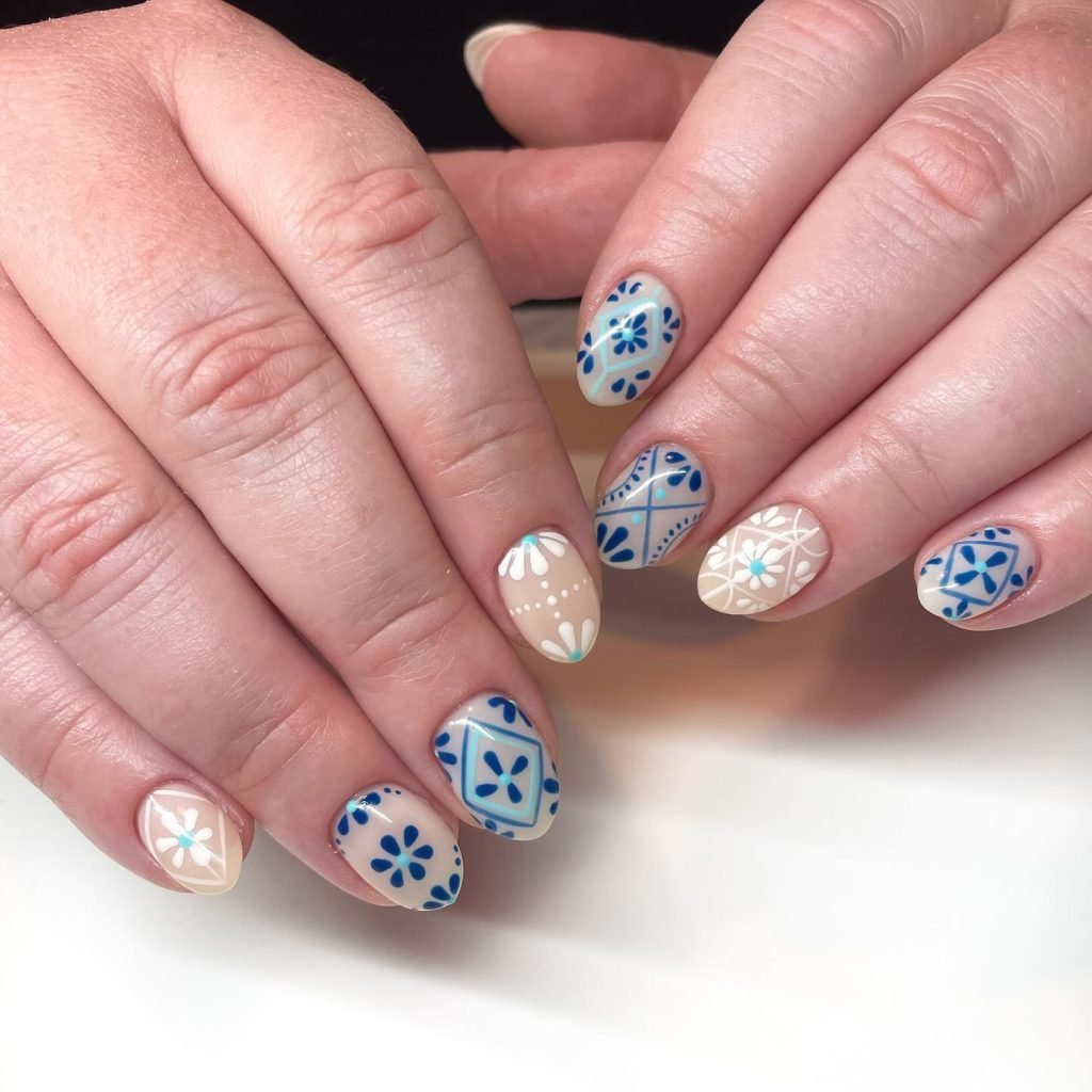 Hands displaying intricate blue and white nail art with floral and geometric patterns on nude nail polish.
