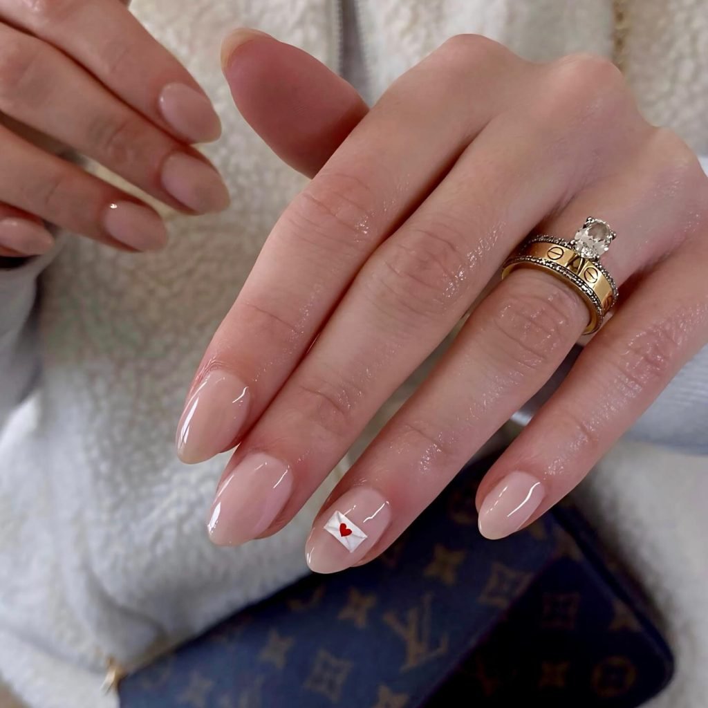 Close-up of a hand with nude manicure, one nail featuring a small heart design, and gold rings on the fingers.