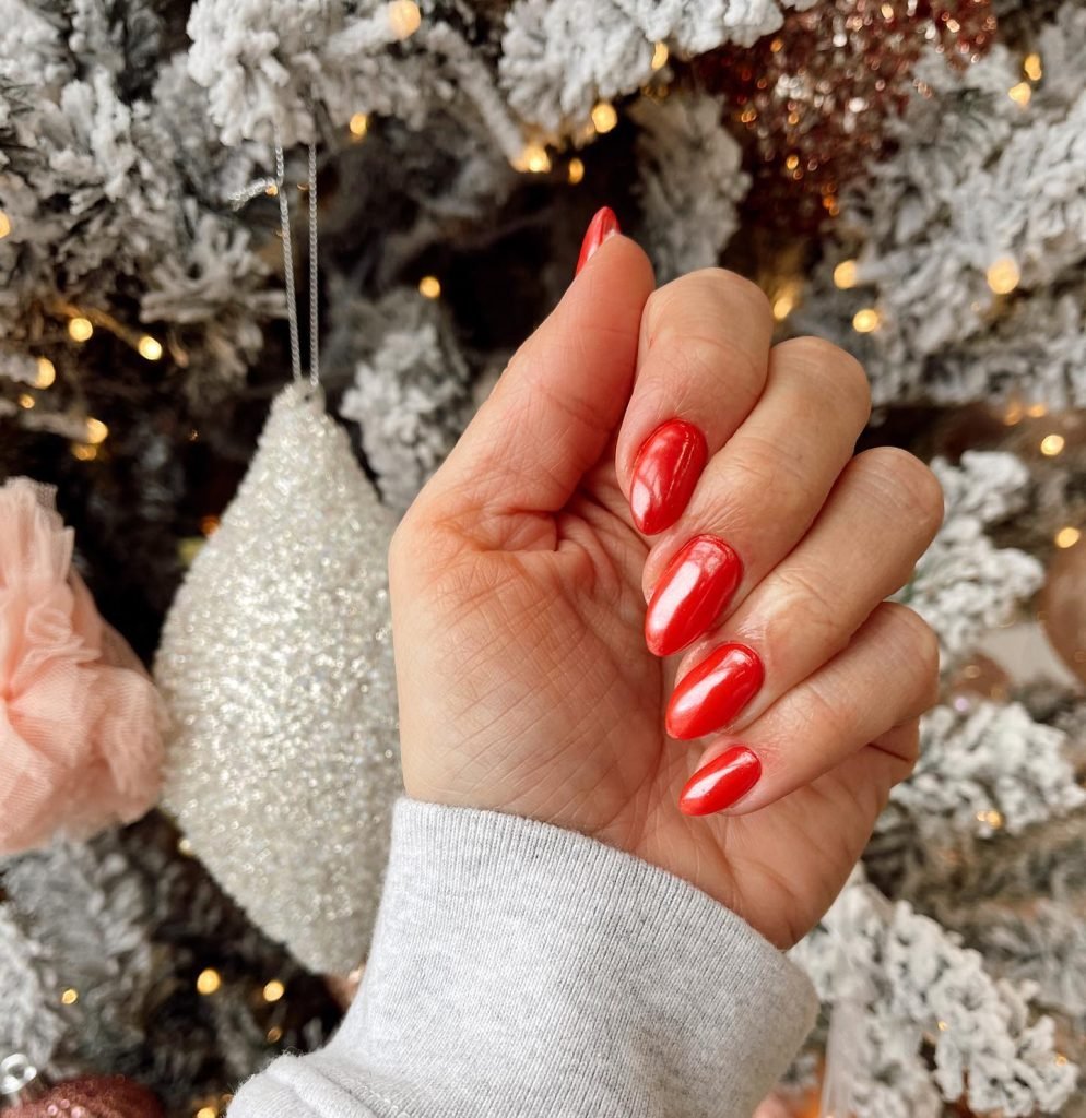 Close-up of manicured hand with red nails, holding ornament near a snow-frosted, lit Christmas tree. Festive nail art.