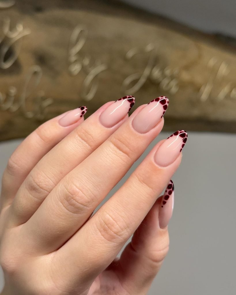Elegant nude nails with black polka dot design on tips, showcasing a stylish and modern manicure.