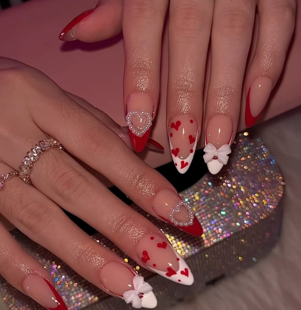 Valentine-themed nail art with red tips, heart accents, pearls, and bows, showcased on manicured hands with rings.
