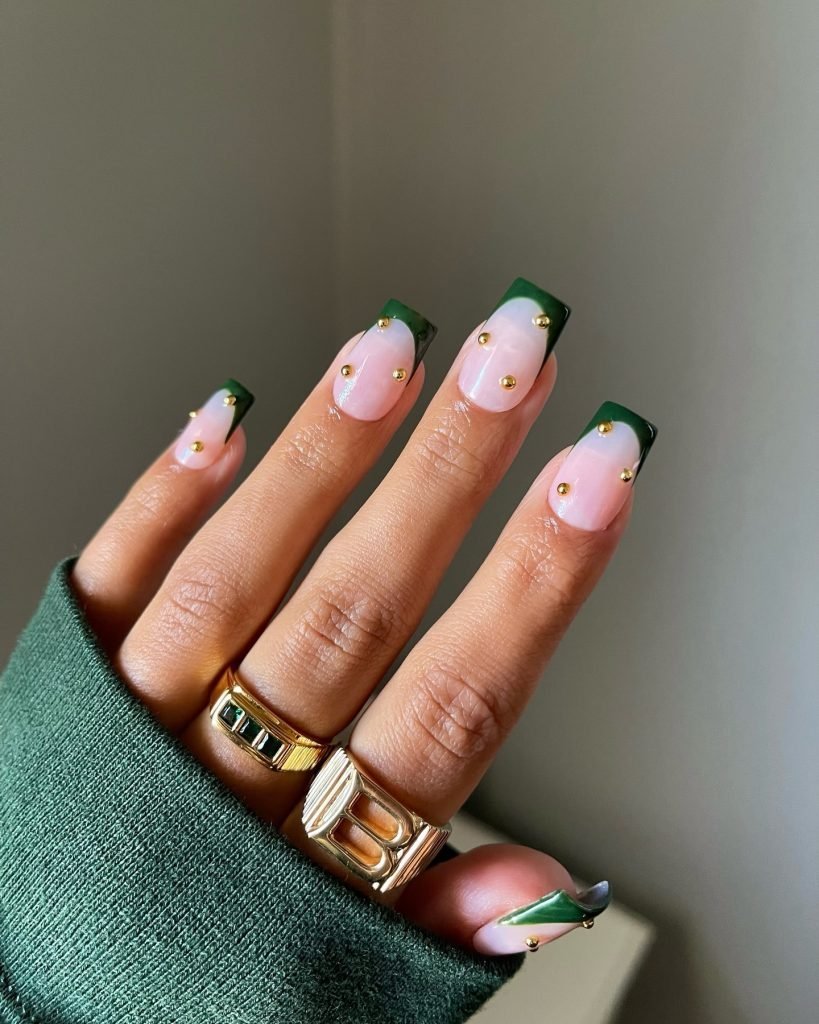 Stylish nails with green French tips and gold studs, complemented by gold rings on fingers wearing a green sweater.