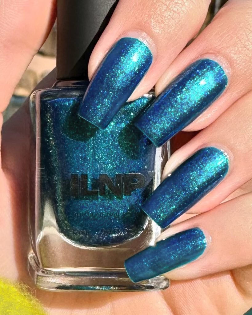 Metallic blue nail polish on fingernails, shimmering in sunlight, next to a nail polish bottle.