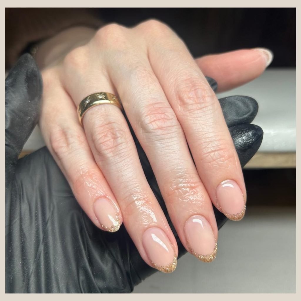 Elegant nude manicure with gold glitter tips on short nails, worn with a gold ring, showcasing professional nail art.