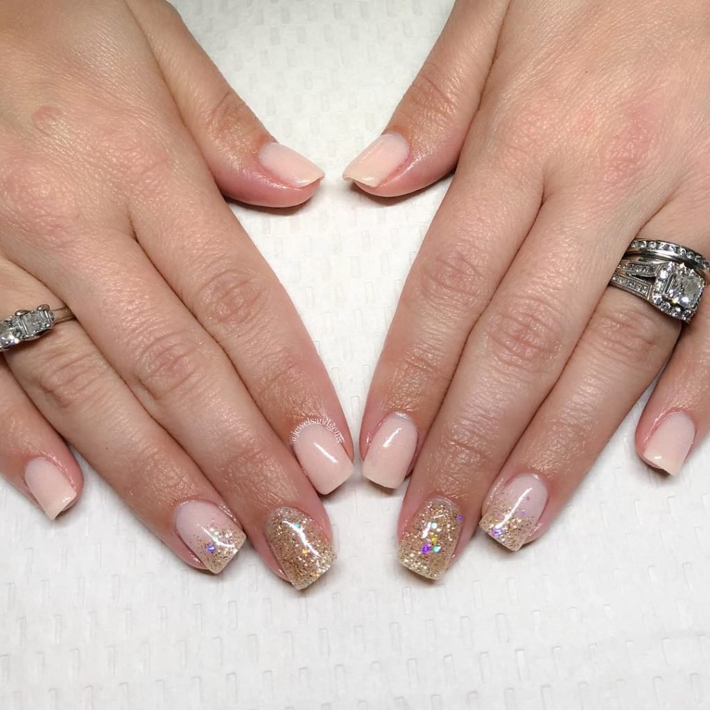 Elegant nude and gold glitter manicure on hands with rings, displayed on a textured white background.