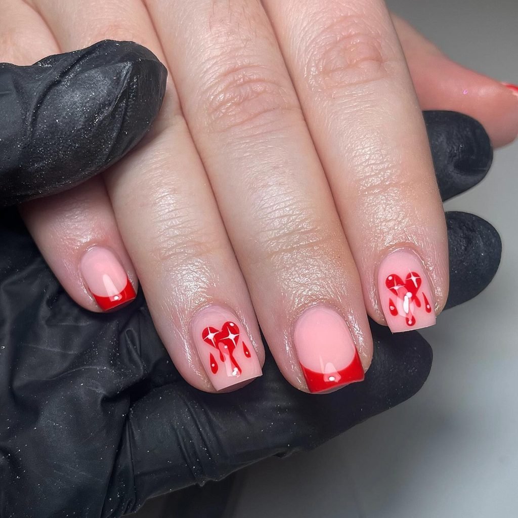 Red French tip nails with heart and drip art on short nails, held by gloved hand.