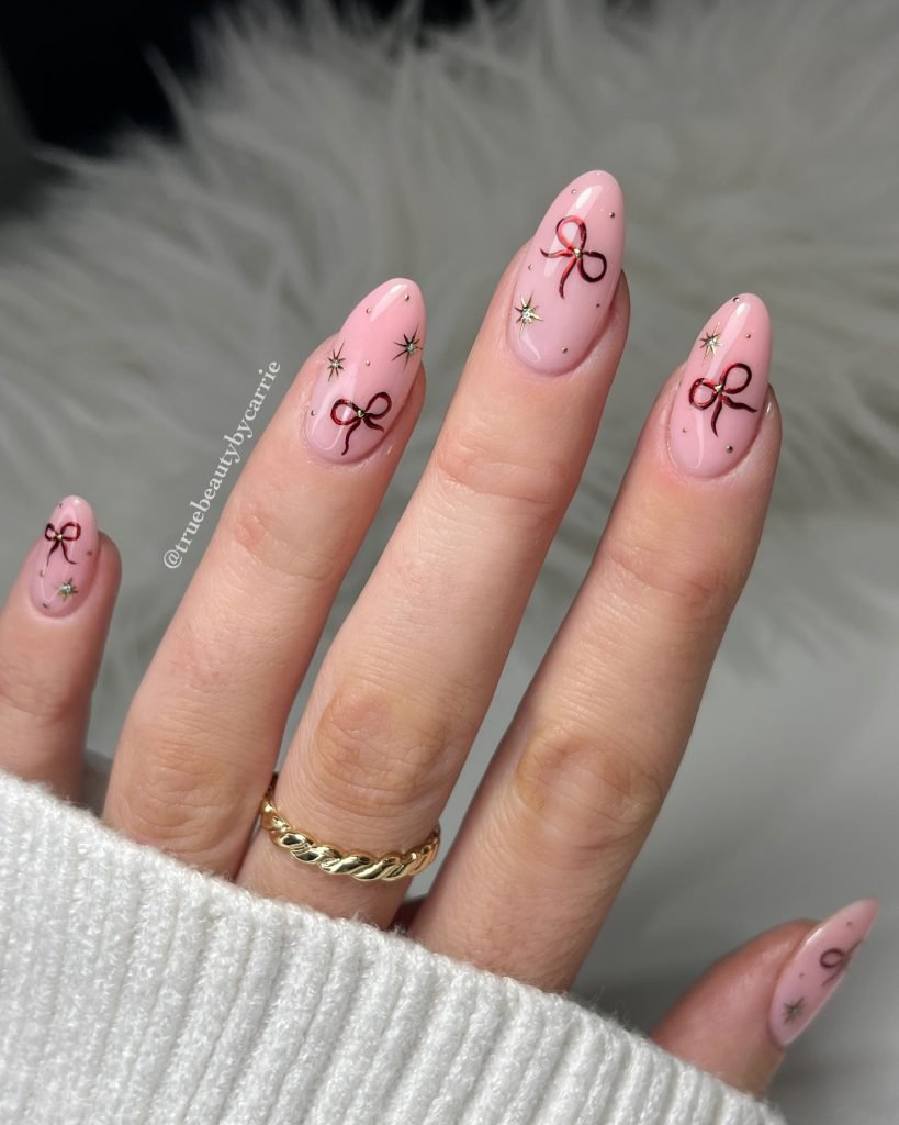 Pink almond nails with red ribbon and star designs, gold ring on finger, cozy white sweater background.