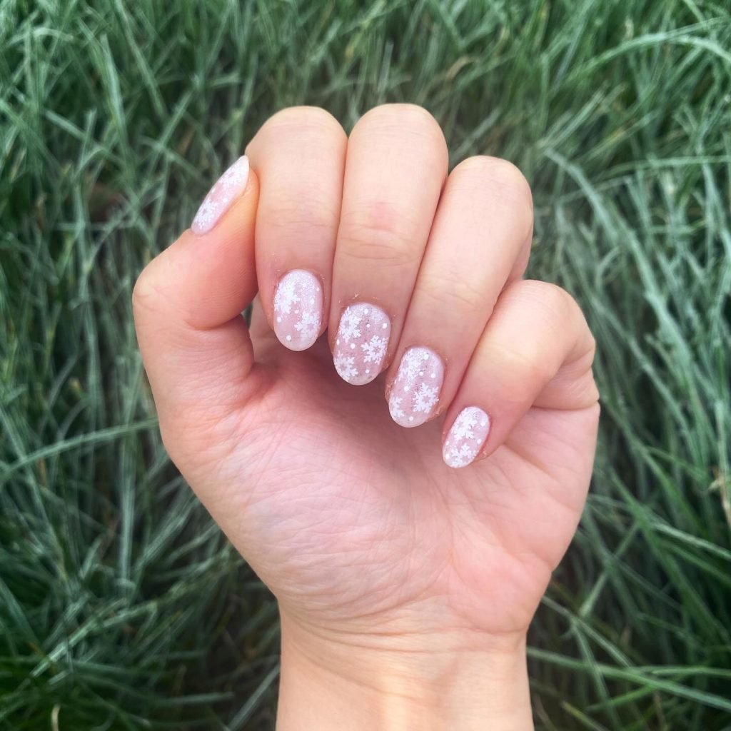 Hand with pink nails decorated with white snowflakes, held against green grass background.