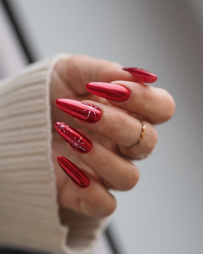 Red metallic nails with intricate white nail art design, close-up, wearing a ring and cozy sweater sleeve.
