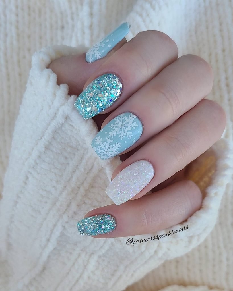 Blue and white winter-themed nails with snowflake patterns and glittery accents against a cozy sweater.