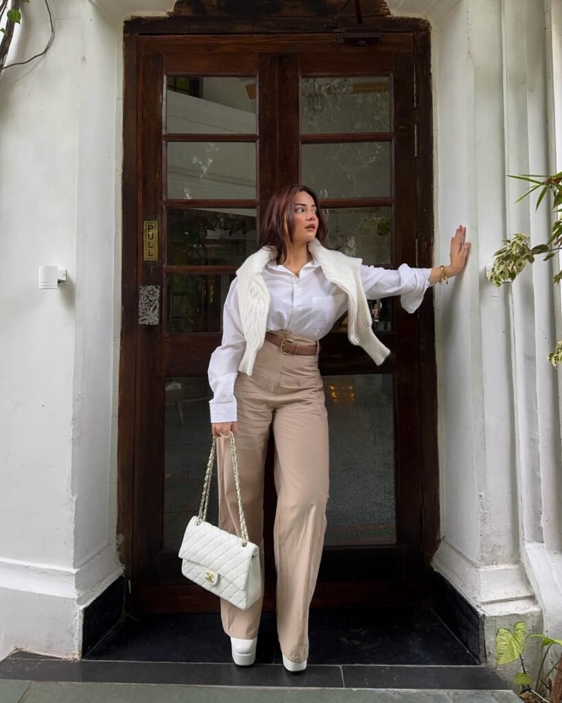Stylish woman in white blouse and beige pants with a handbag, standing by a wooden door.