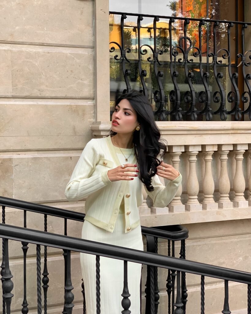 Woman in cream outfit poses by stylish railing and stone building, embracing elegant and modern fashion vibes.