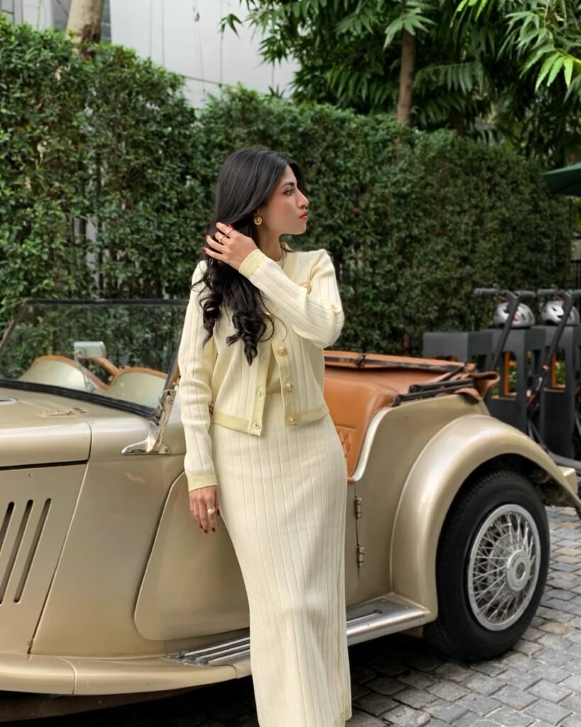 Woman in beige outfit standing by vintage car, surrounded by lush greenery.