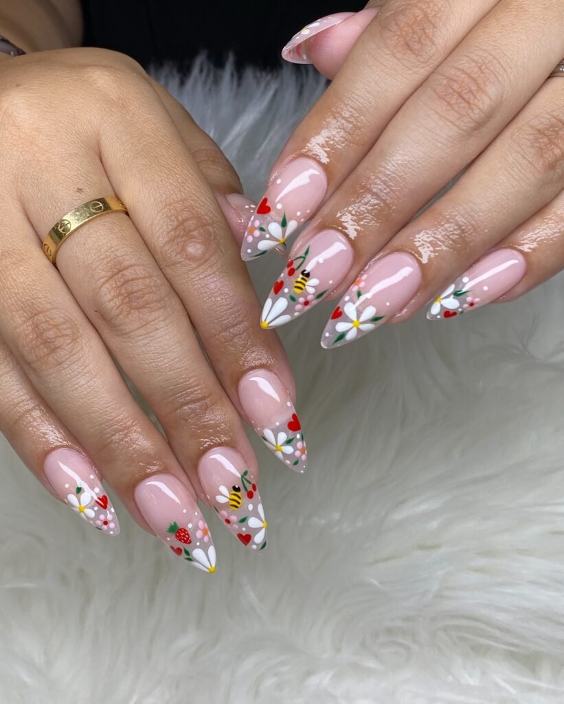 Colorful spring nail art with daisies, bees, hearts, and strawberries on almond-shaped nails.
