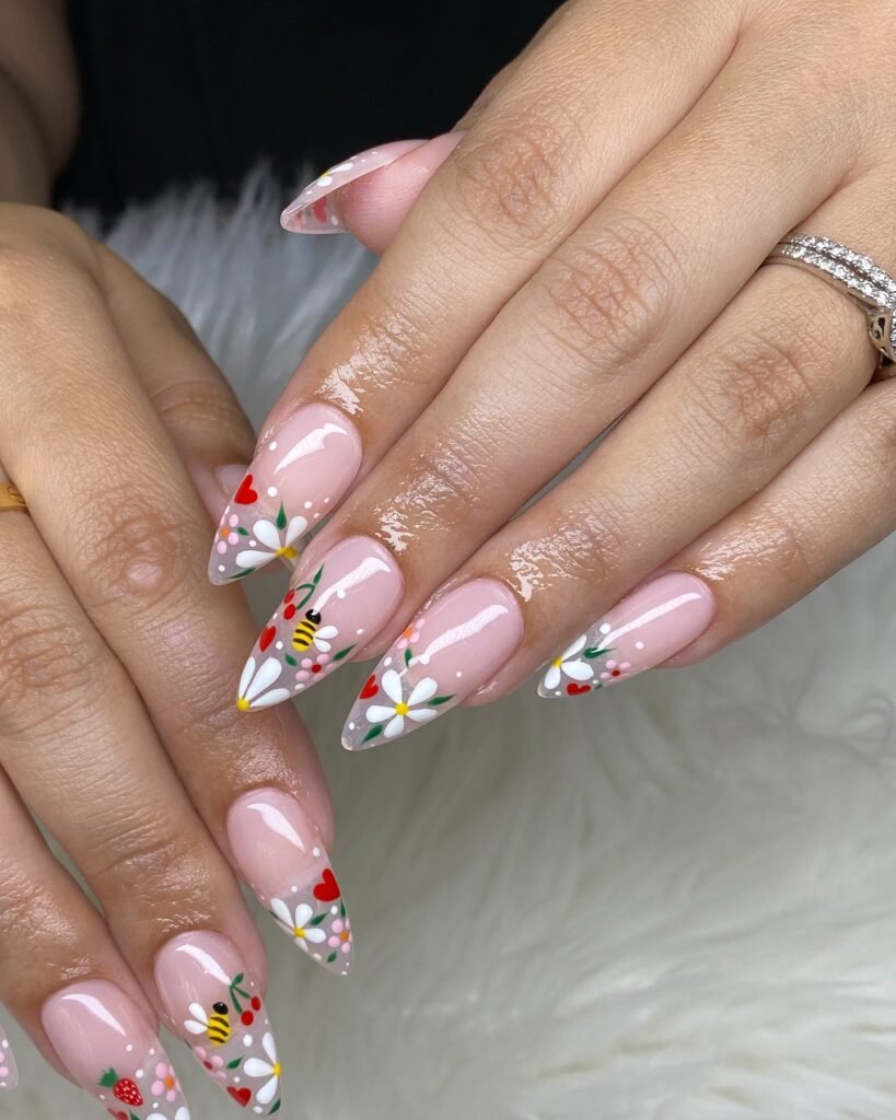 Colorful floral and bee nail art design on pointed nails, featuring daisies, strawberries, and hearts.
