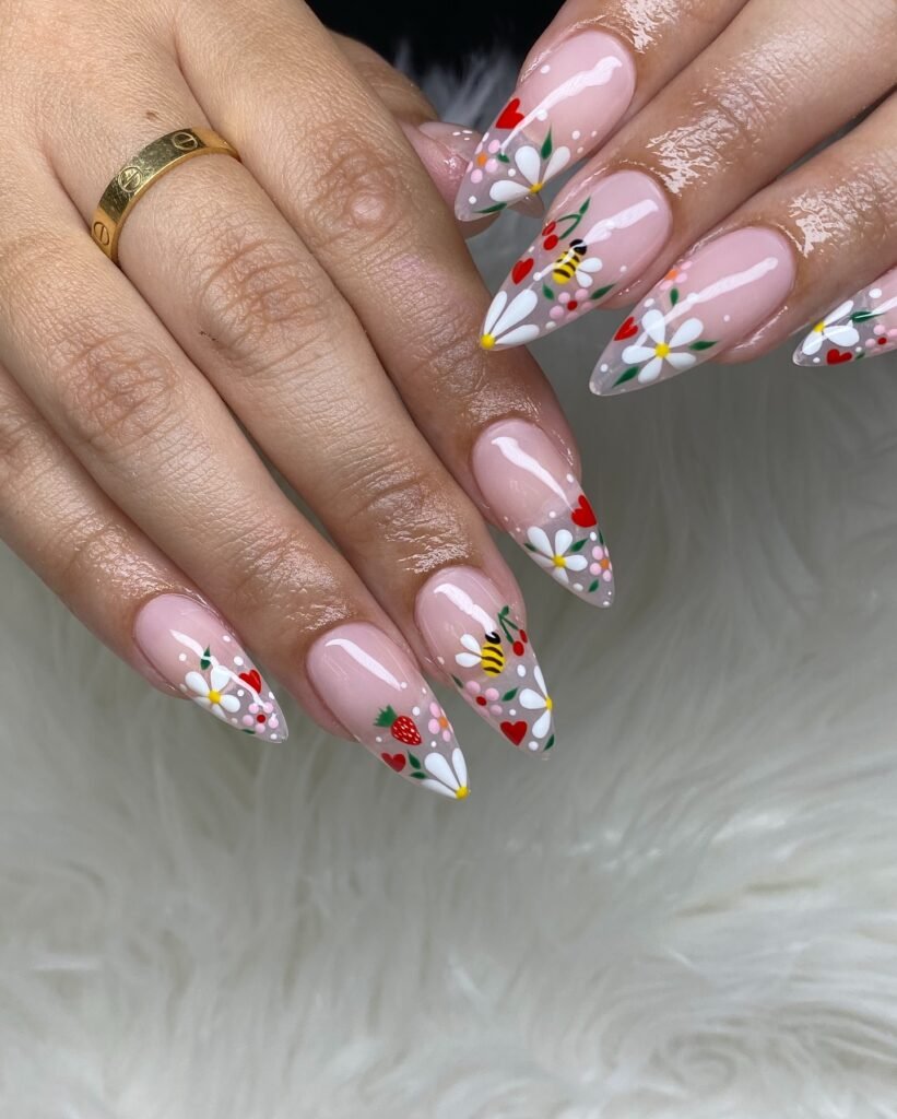Colorful floral nail art with daisies, hearts, bees, and strawberries on long almond-shaped nails, hand on white fur.