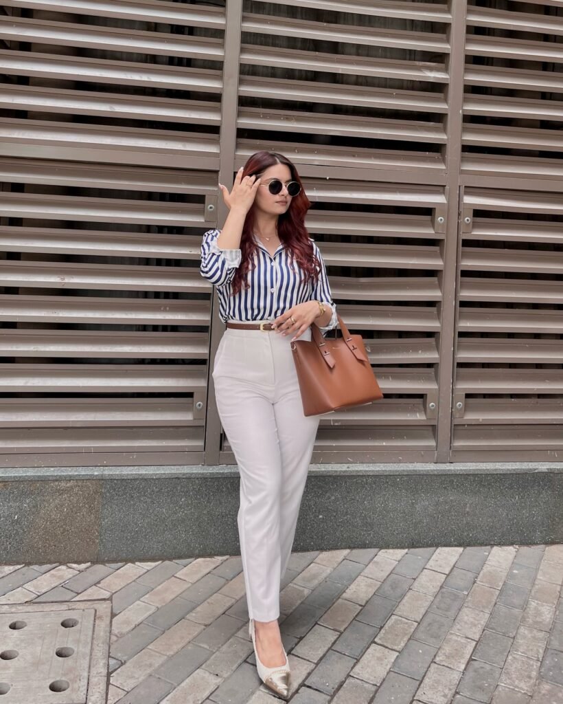 Woman in striped shirt and white pants with brown handbag standing by a modern building wall.