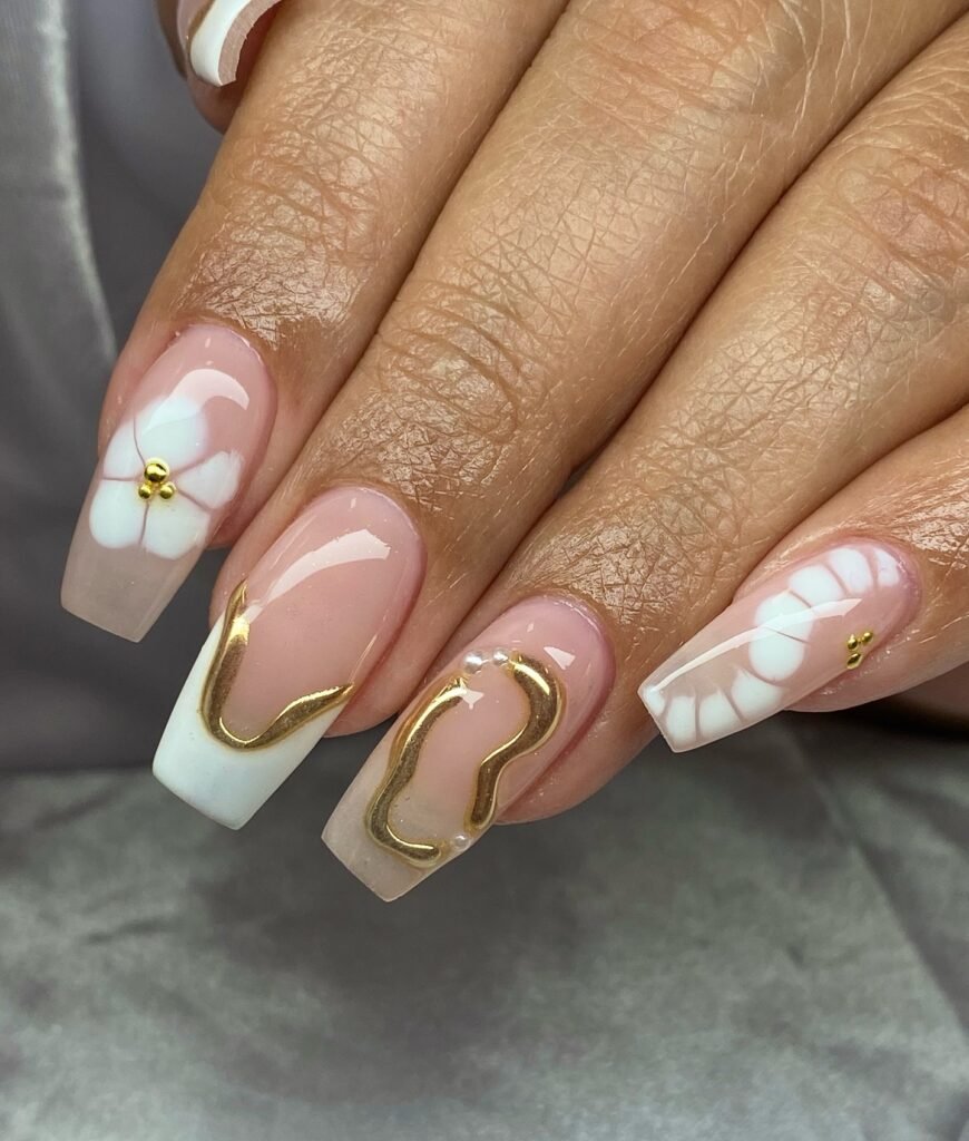 Elegant nude and white nail art with gold accents and floral designs on coffin-shaped nails.