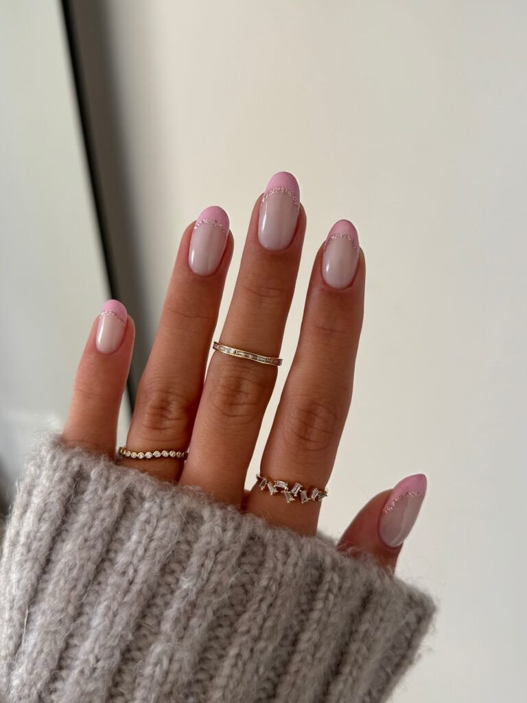 Pink French manicure with silver accents on manicured nails, adorned with gold rings, wearing a cozy gray sweater.
