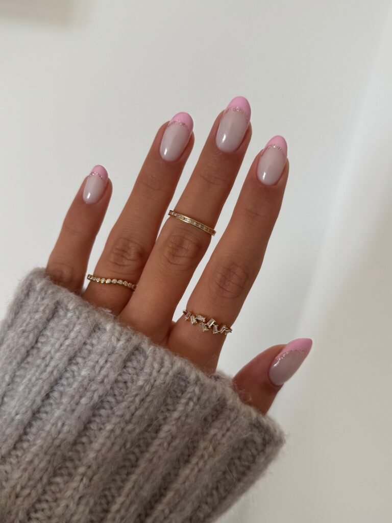 Pink French manicure with gold rings on a hand wearing a gray sweater sleeve.