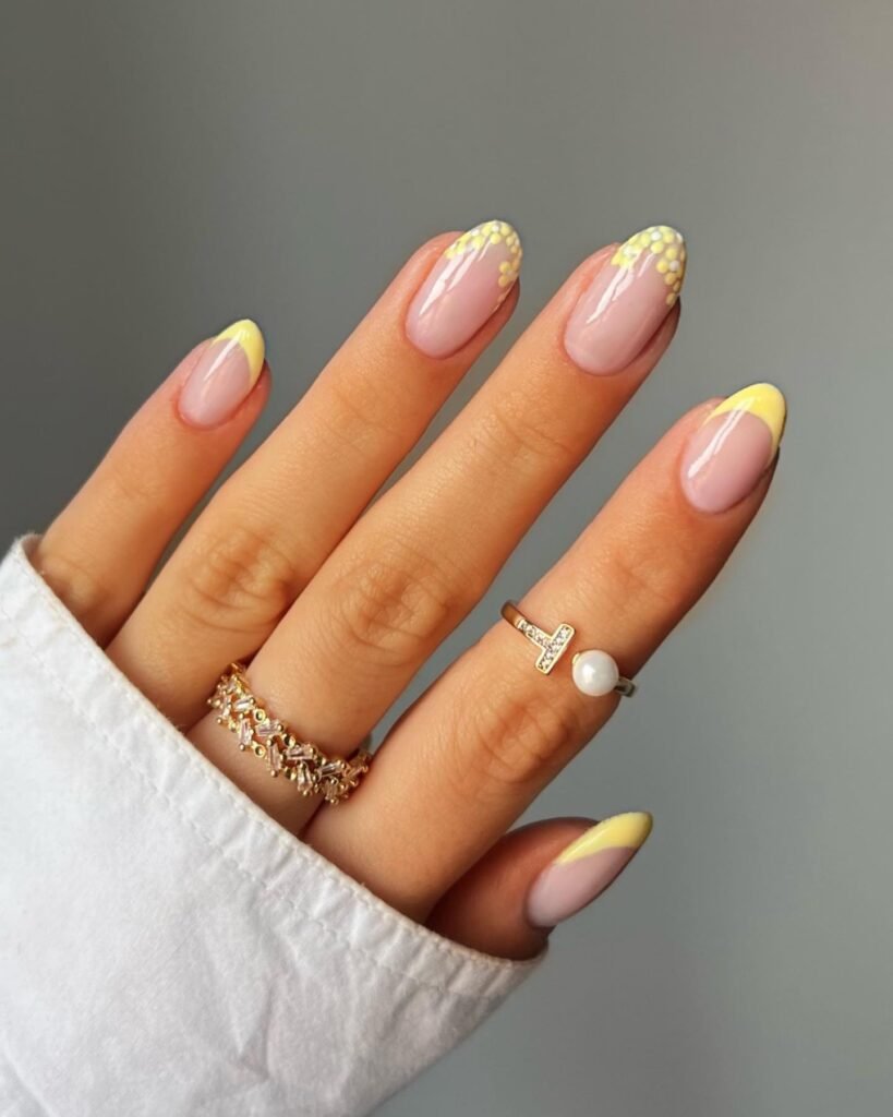 Stylish yellow floral nail art with gold and pearl rings on a hand wearing a white shirt.