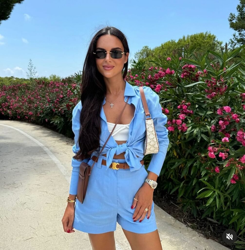 Stylish woman in blue outfit and sunglasses posing near blooming flowers on a sunny day.