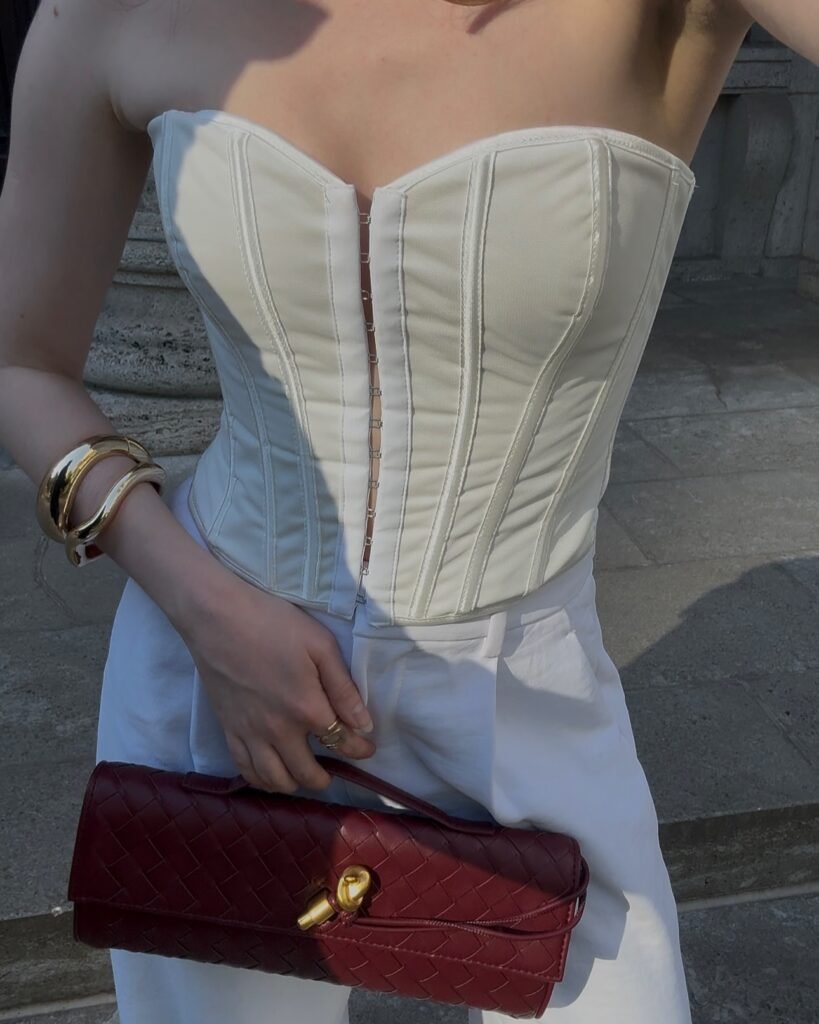 Woman in white corset top with gold bangles holding a red woven clutch.