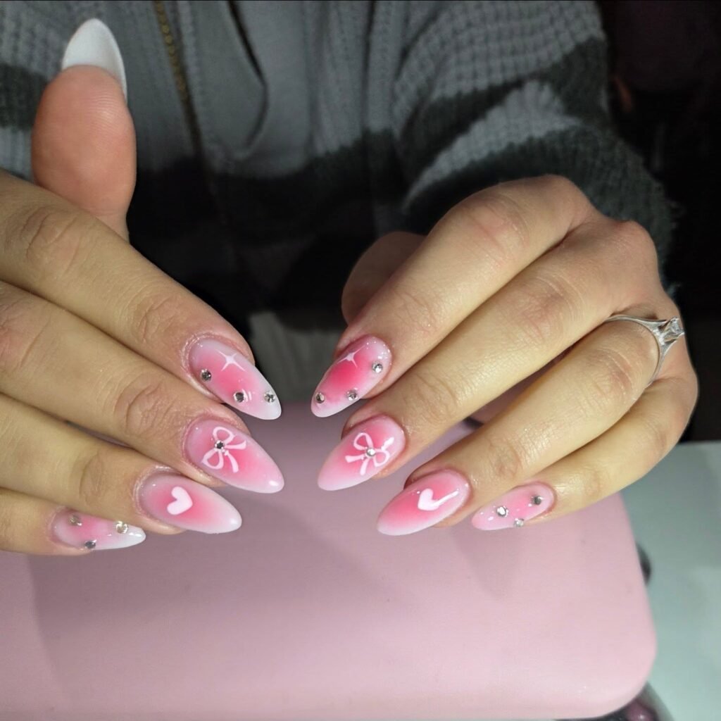Pink nail art with bows and hearts on stiletto-shaped nails, adorned with rhinestones for a stylish look.