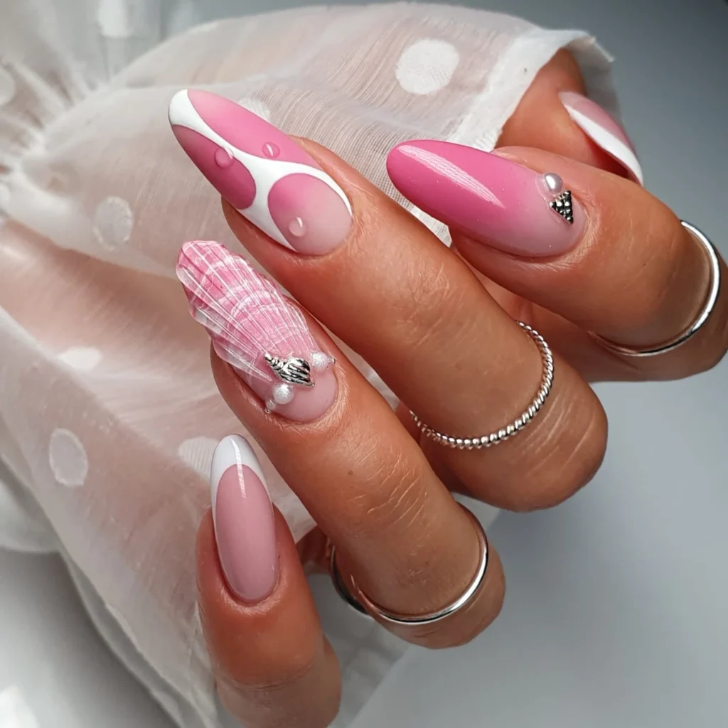 Pink and white artistic nail design with 3D shell accents and glossy finish on manicured hands wearing silver rings.