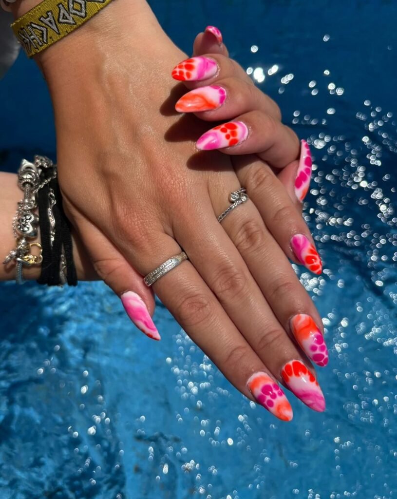 Bright pink and orange nail art with bold patterns, sparkling against a blue water background.