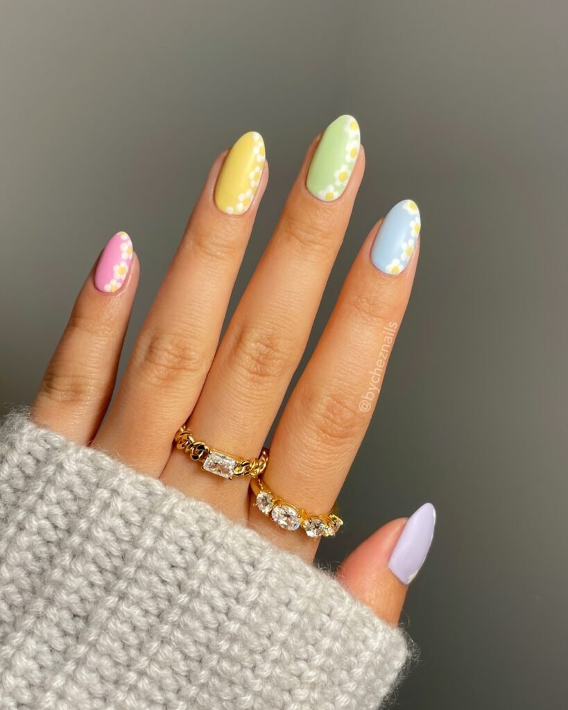 Colorful daisy nail art with pastel shades on almond-shaped nails, accessorized with gold and diamond rings.