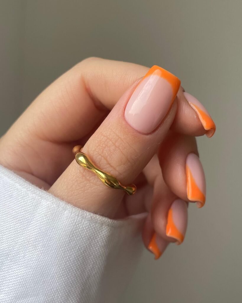 Hand with orange French tip manicure and gold twisted ring.