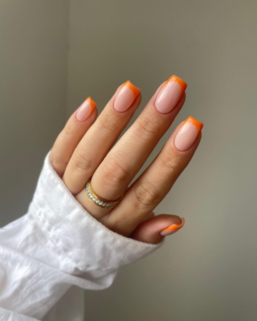 Stylish hand with nude nails and orange French tips, wearing a gold ring, white shirt sleeve visible.